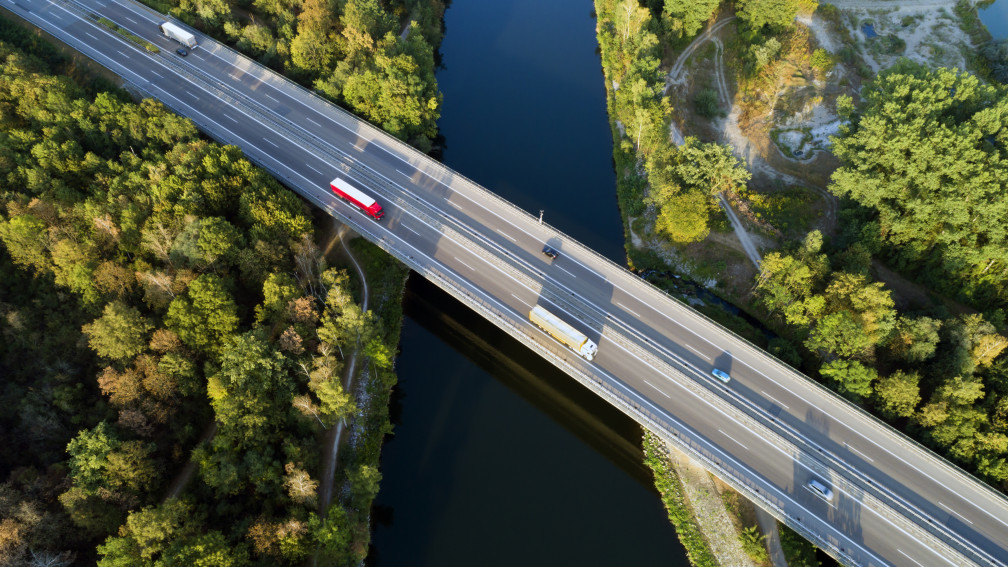 Lastwagen fahren über Brücke; darunter Wälder und ein Fluss