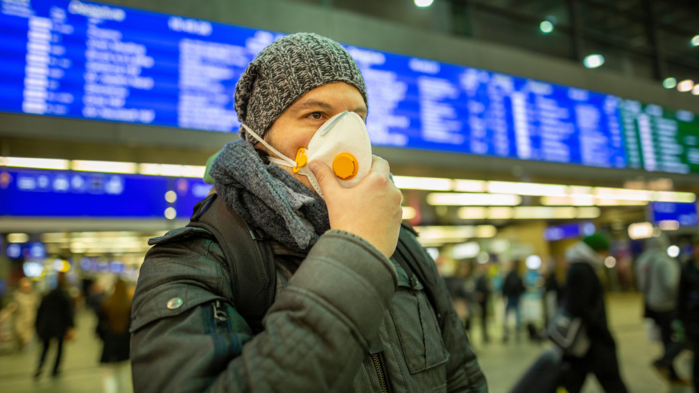 Mann mit Schutzmaske steht am Bahnhof vor einer digitalen Anzeigetafel