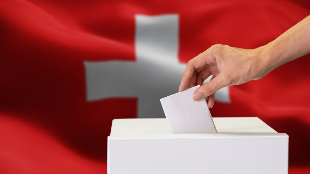 Hand wirft Wahlzettel in Wahlbox ein, im Hintergrund eine Schweizerflagge