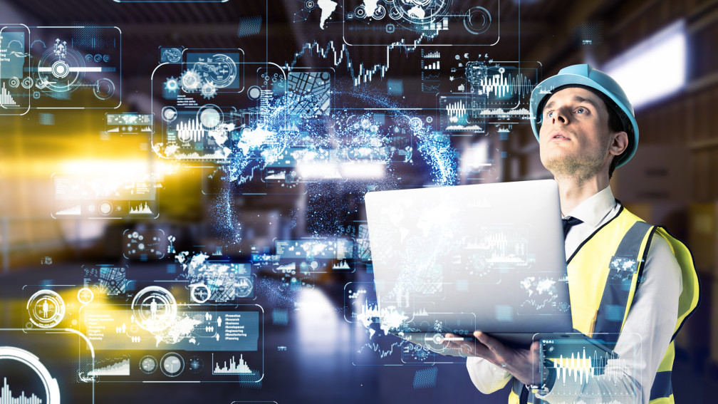 Mann in einem Industriebetrieb mit Laptop und digitaler Projektion vor sich
