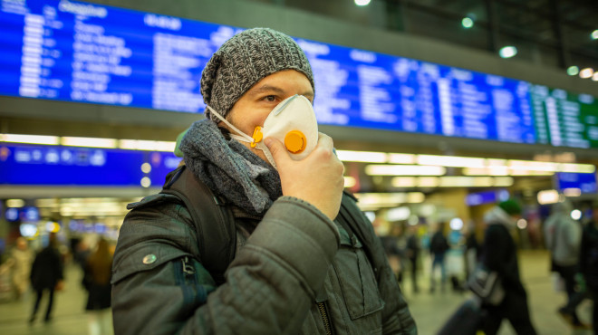 Mann mit Schutzmaske steht am Bahnhof vor einer digitalen Anzeigetafel