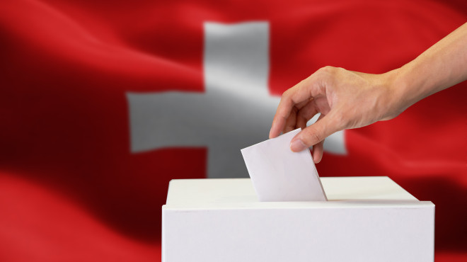 Hand wirft Wahlzettel in Wahlbox ein, im Hintergrund eine Schweizerflagge