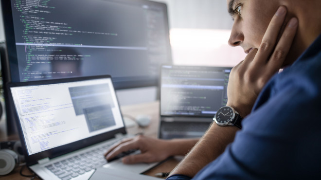 Mann sitzt vor drei Computer Screens und codiert