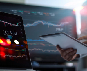 2 Computerbildschirme mit Statistiken darauf und eine Hand mit IPad ein Finger zeigt darauf